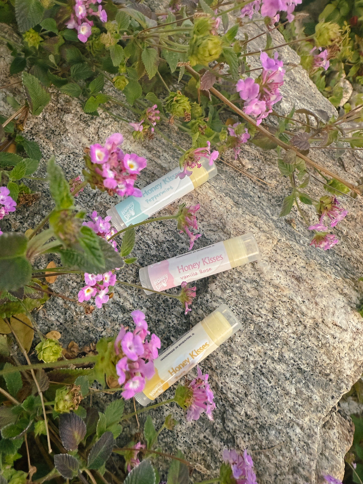 Honey Kiss lip balms sit on a rock surrounded by flowers