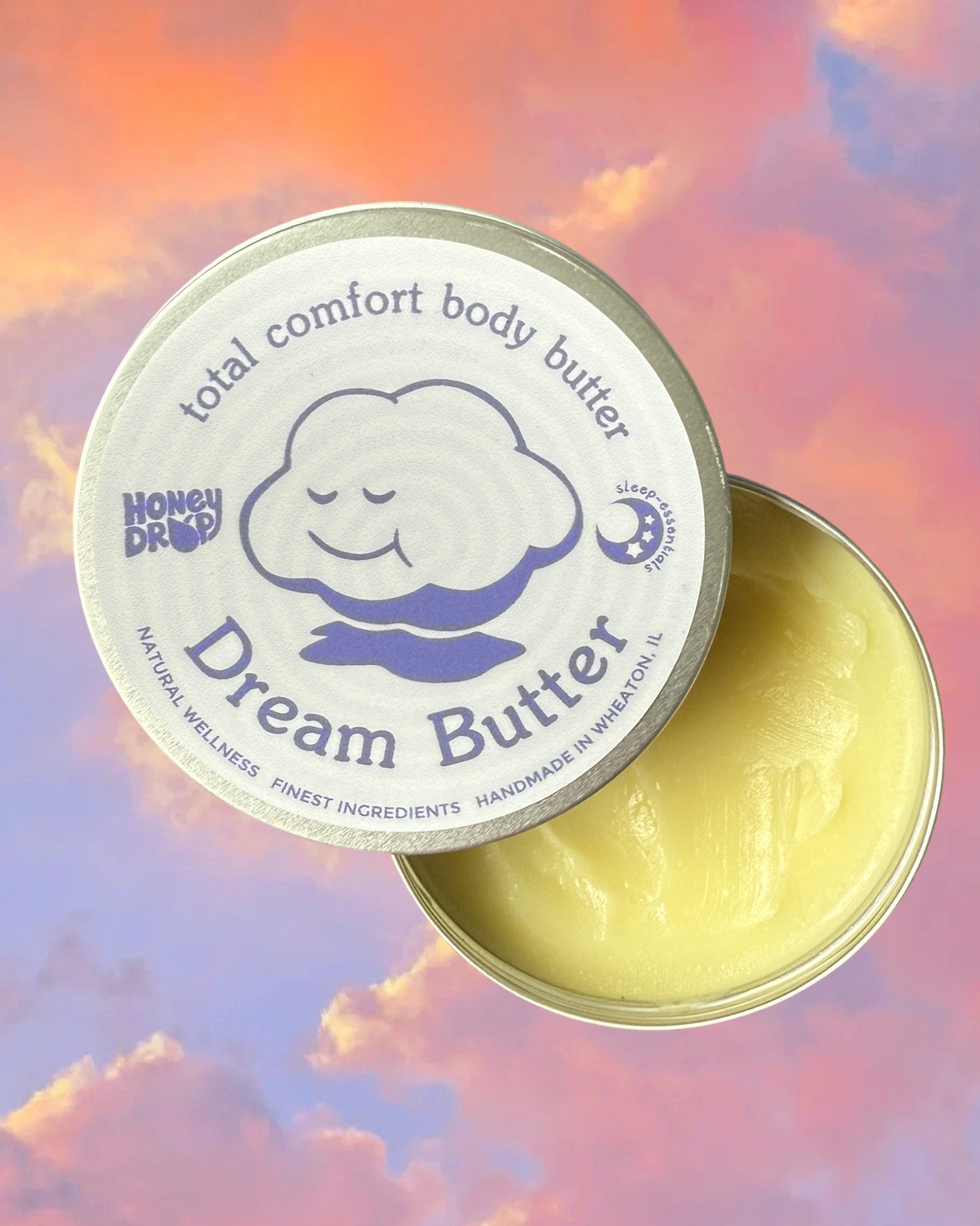 An open round metal tin of Honey Drop Dream Butter body butter, showing the pale yellow balm inside, with the lid placed beside it. The lid label is white with blue text and a smiling cloud illustration, set against a pastel pink, orange, and blue sky background.