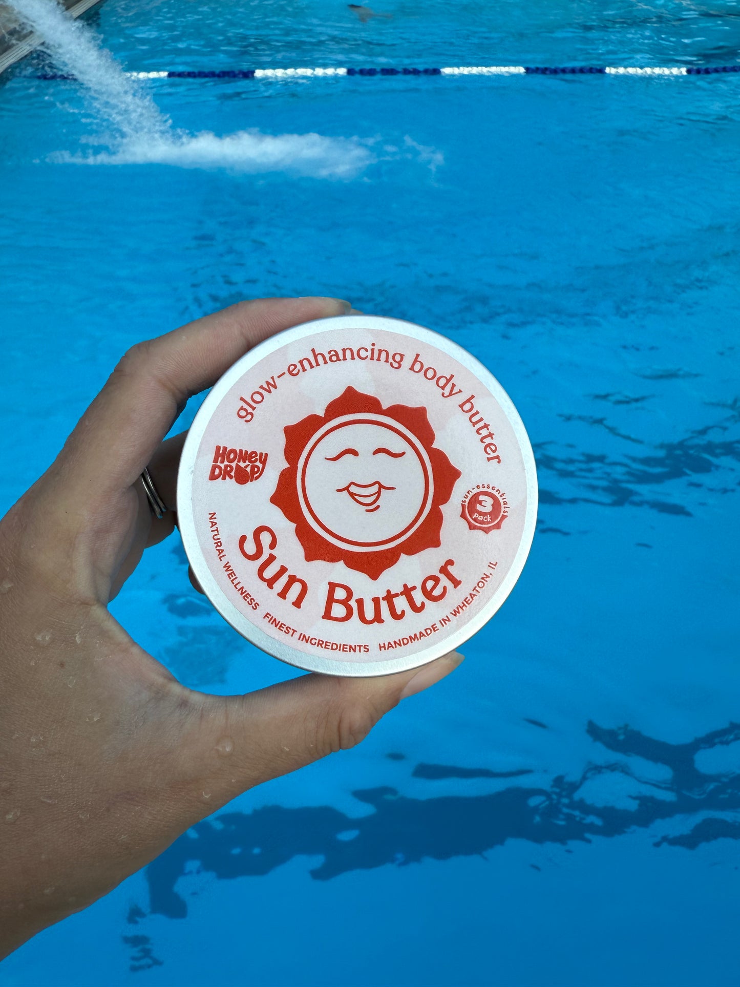 A hand holding a round white tub of 'Sun Butter' body butter in front of a bright blue swimming pool background.