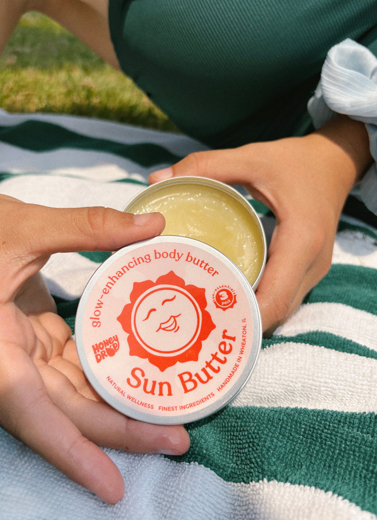 A person outdoors holds an open round tin of Sun Butter body butter over a striped green and white blanket. The lid shows the brand name Sun Butter with a red sun logo, and the open tin reveals a pale yellow balm inside.
