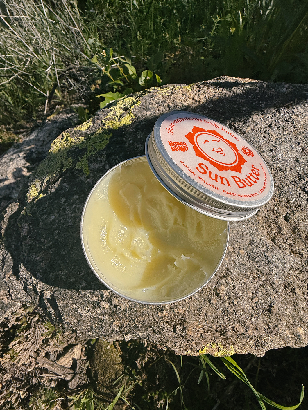 An open round metal tin of Sun Butter balm with pale yellow cream inside, placed on a large rock outdoors with grass and sunlight visible. The lid is resting beside the tin and shows the Sun Butter branding.