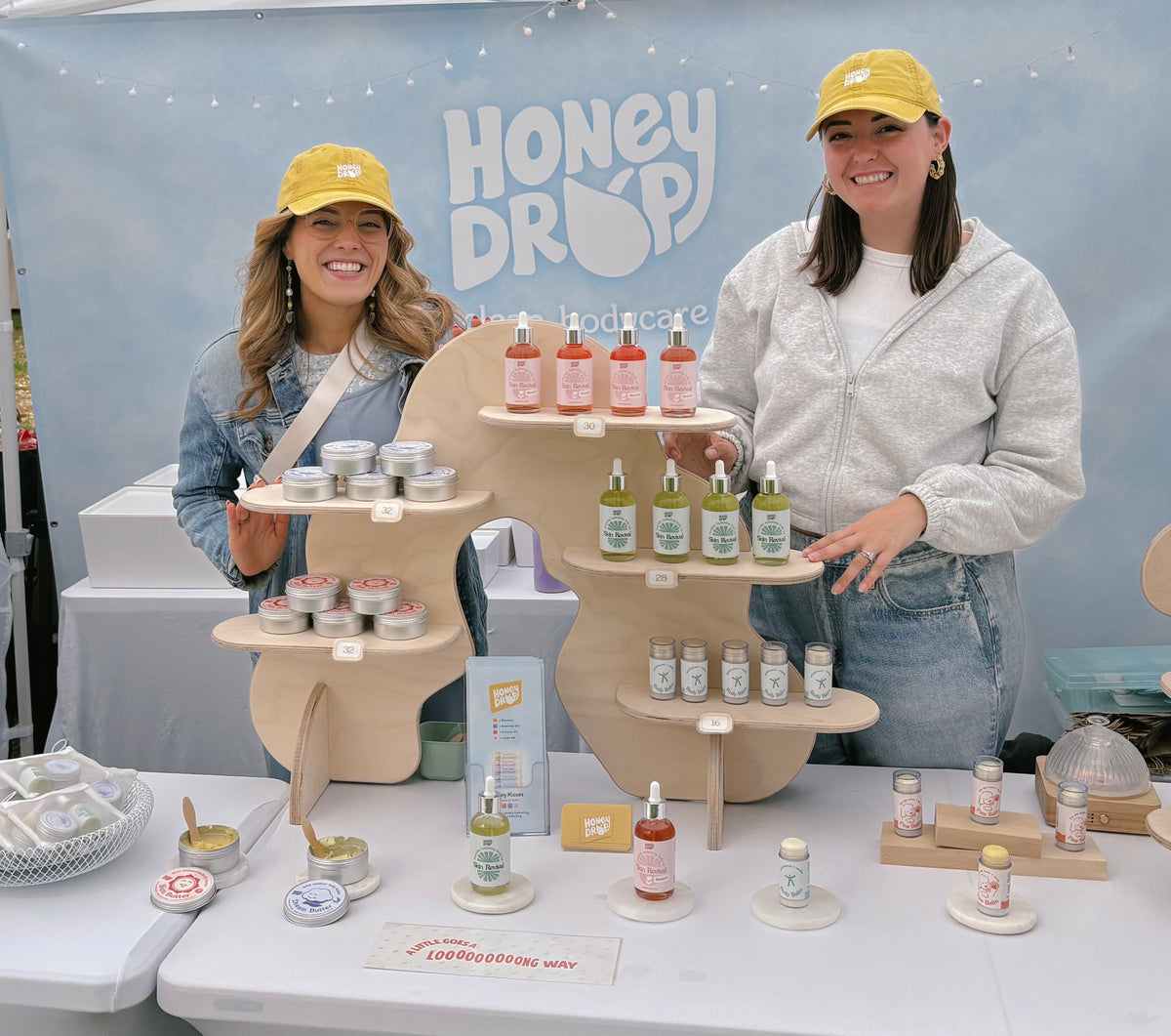 Two women wearing yellow Honey Drop hats stand behind a display table filled with skincare products and wooden tiered shelves. A large Honey Drop backdrop is visible behind them.