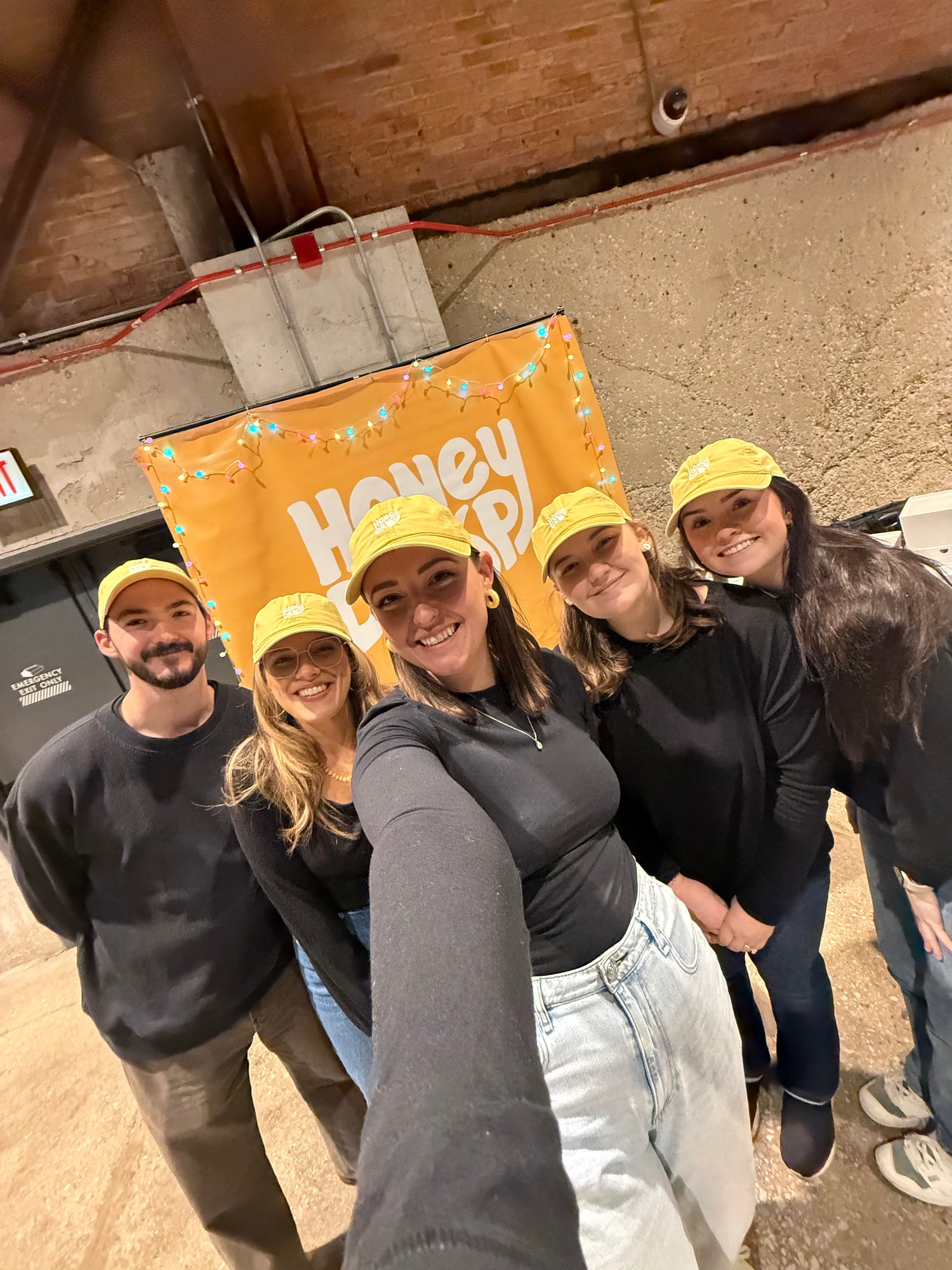 Five people wearing matching black long-sleeve shirts and yellow baseball caps pose for a selfie in front of an orange banner that reads 'Honey Drop'.