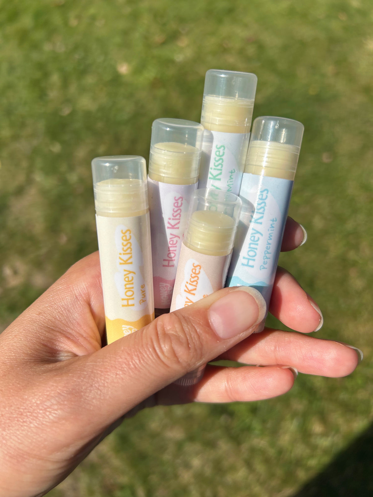 A hand holding five tubes of lip balm outdoors on a grassy lawn. The lip balms are labeled 'Honey Kisses' and come in yellow, pink, and blue flavors.