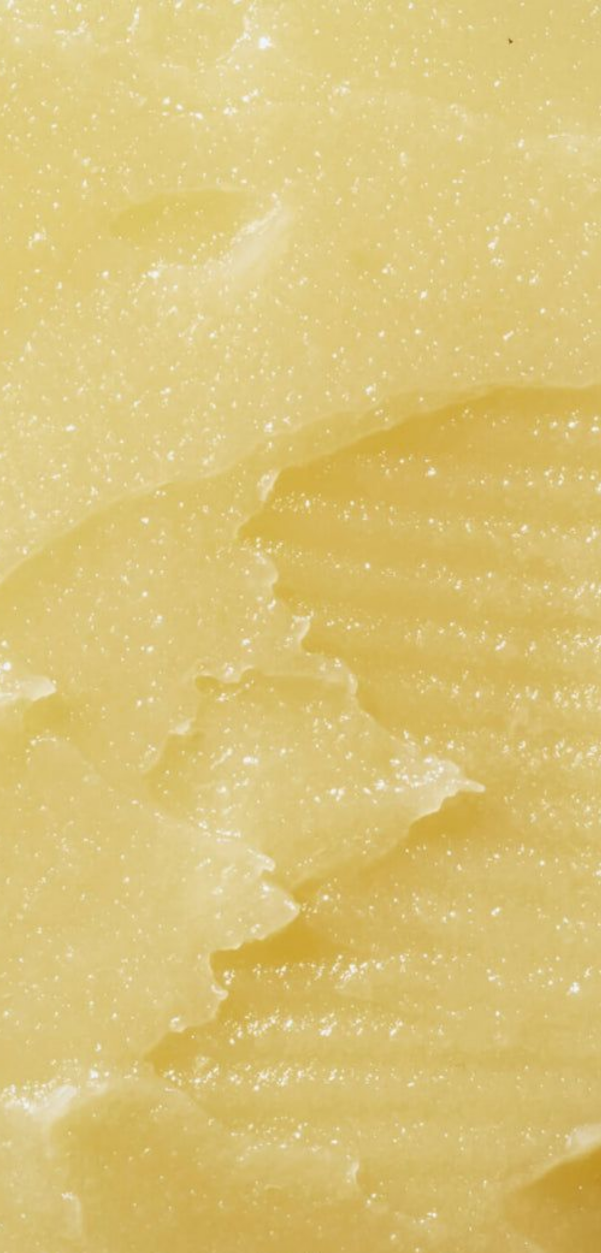 A close-up texture shot of a pale yellow, creamy solid mango butter with a slightly glossy, granular surface and a ribbed pattern on the right side.