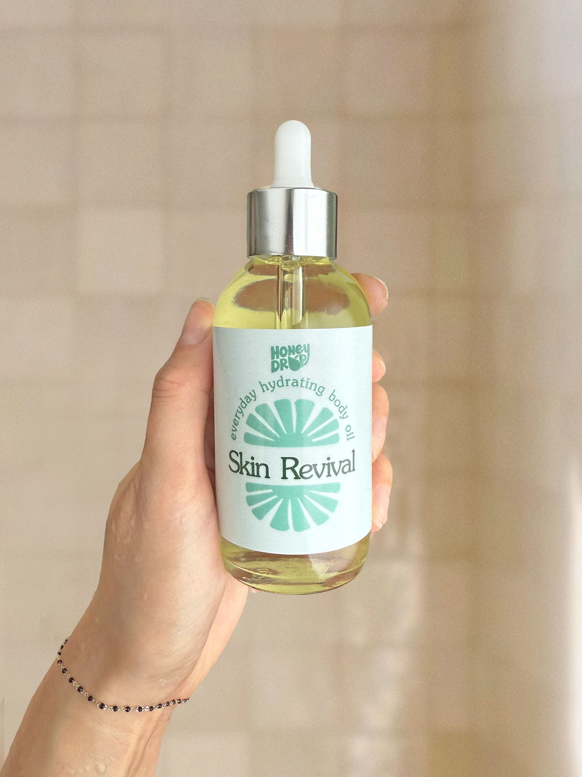 A hand holding a glass dropper bottle of Home Based Body Oil Skin Revival with a white label and silver cap, set against a soft beige tiled background.