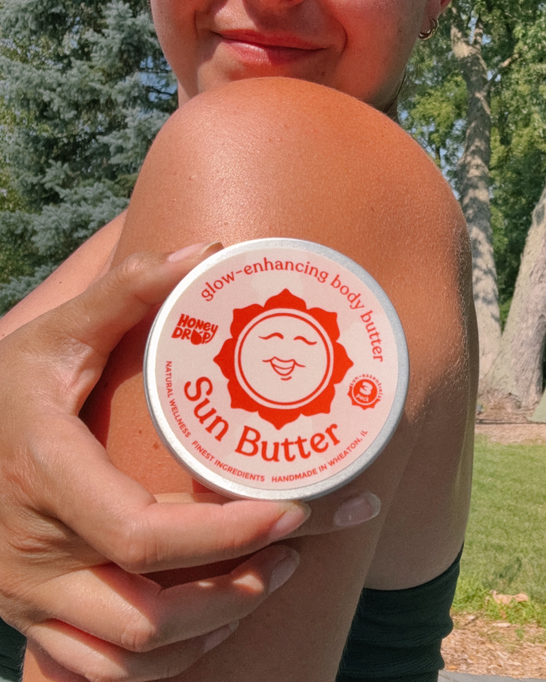 A person outdoors holds a round tin of Nowen Doody Glow-Enhancing Body Butter on their shoulder. The product packaging is white with a prominent red sun logo and text.