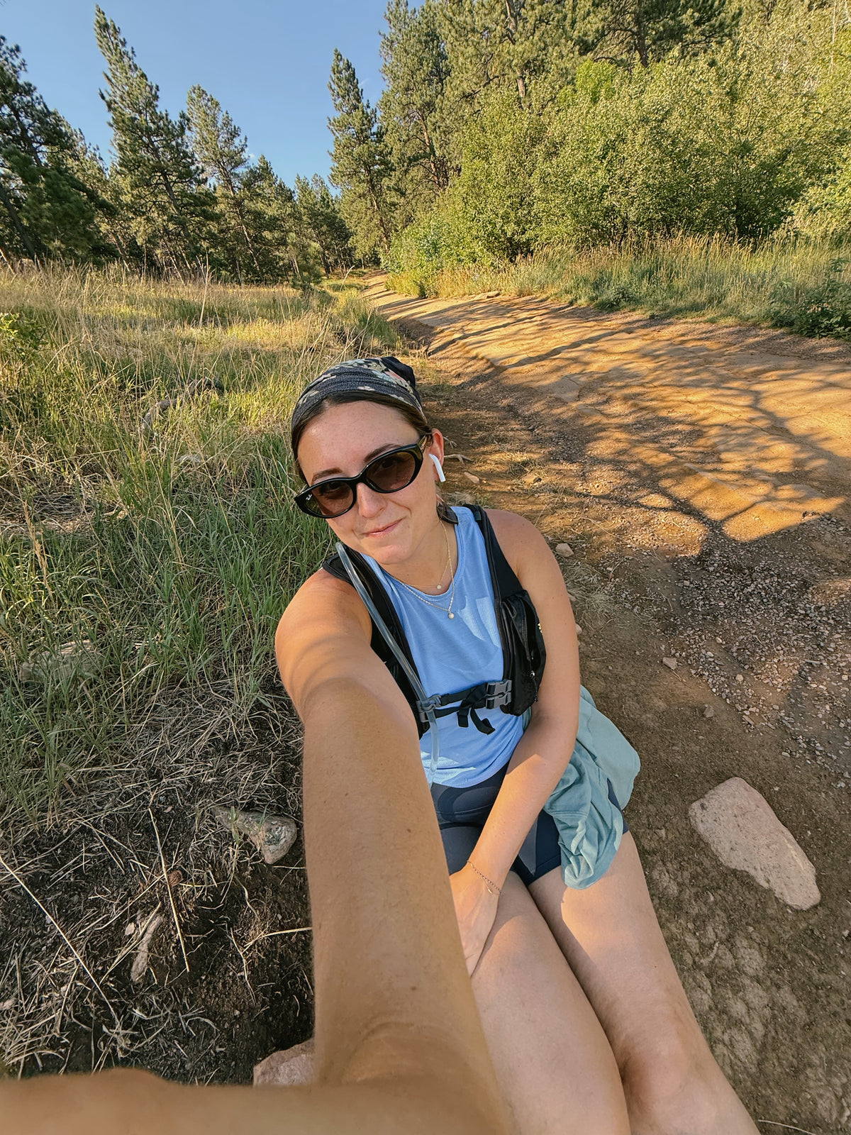 A woman taking a selfie on a dirt hiking trail in a grassy forest clearing, wearing a light blue tank top, dark shorts, sunglasses, and a hydration pack.