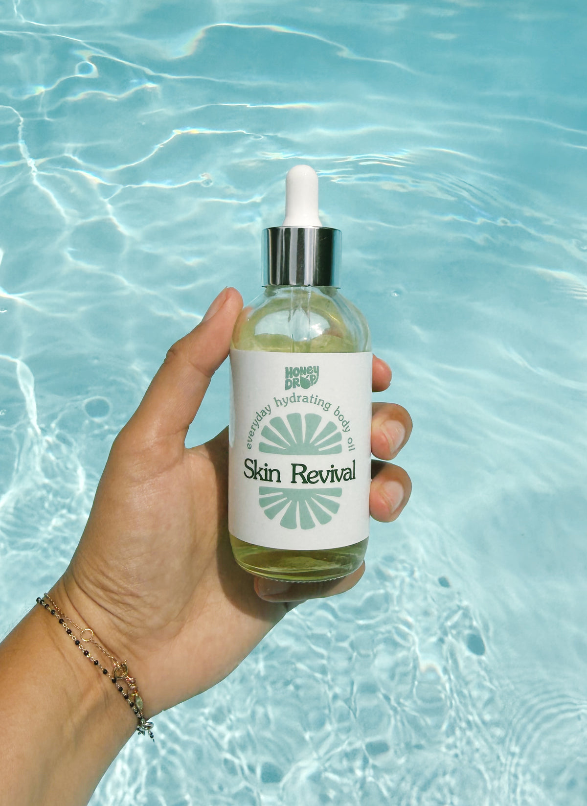 A hand holds a clear dropper bottle of Honey Bee Body Oil labeled 'Skin Revival' against a bright blue swimming pool water background.